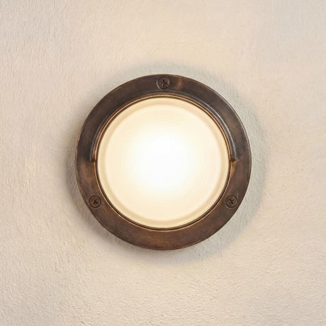 Walkway Eyelid Outdoor Wall Light in antique brass, round solid brass body with eyelid hood over a frosted glass lens, screw fixings visible, mounted on a plain beige wall.