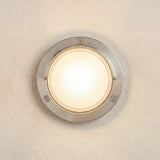 Walkway Eyelid outdoor wall light in satin nickel, round brass fixture with frosted glass diffuser and curved eyelid hood, illuminated, IP65 rated, mounted on a light wall.
