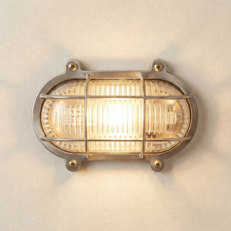 Merchant Flush Oval Bulkhead Light in satin nickel, showing the ribbed glass diffuser under a barred cage with visible mounting screws, lit against a plain wall.