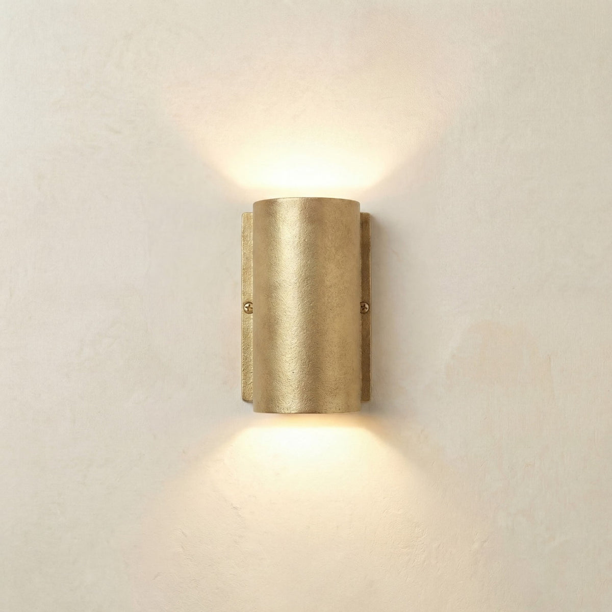 Pharos Compact Outdoor Up & Down Wall Light in raw brass, a half-cylinder fixture on a slim wall plate with visible screws, casting light up and down on a plain light background.