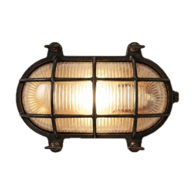 Horatio oval bulkhead light in blackened brass, with ribbed glass behind a grid-barred brass cage and corner fixing lugs, lit, on a plain white background.