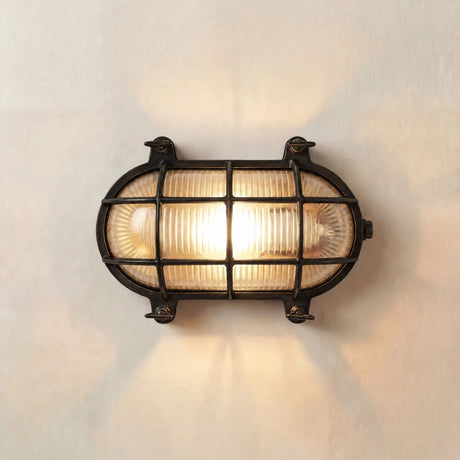 Horatio Oval Bulkhead Light IP64 in blackened brass, lit on a plain wall, showing ribbed glass behind a gridded brass cage with corner fixing lugs.