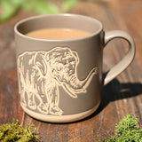 Grey glazed ceramic mug with wax-resist elephant design in fine pale line work, raised trunk near the handle, filled with tea on a wooden surface.