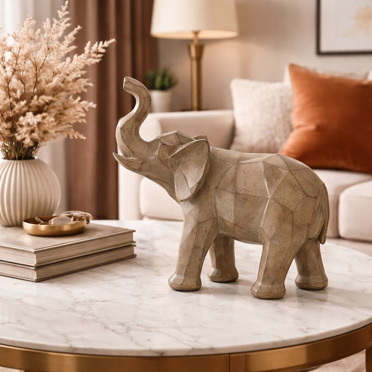 Geometric Playful Elephant Figurine with Raised Trunk in grey, faceted finish, displayed on a marble coffee table in a living room with cushions and dried pampas grass.