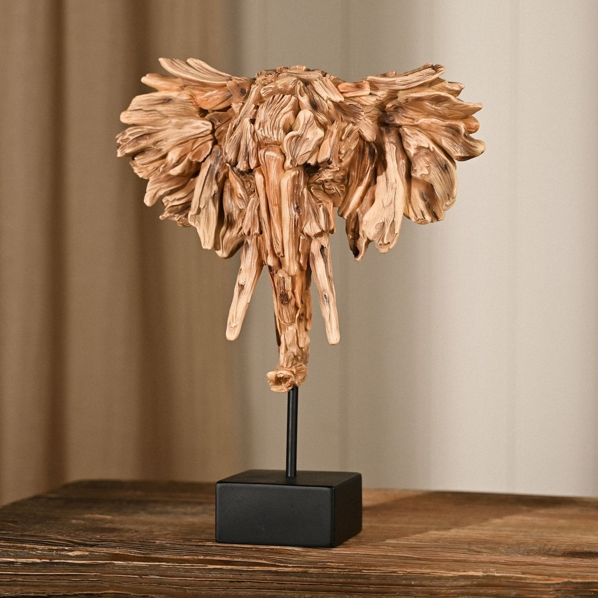 Driftwood Elephant Head Resin Figurine with Stand in brown, featuring fanned ears, downward tusks and curled trunk on a black base, shown on a wooden table.