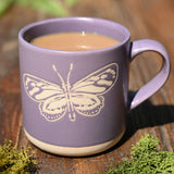 Purple stoneware mug with cream base, featuring a wax resist butterfly design in cream against the lavender glaze, filled with tea and shown outdoors on a wooden surface.