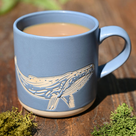Blue stoneware mug with a cream engraved whale design and loop handle, filled with tea, set on a wooden surface with moss.