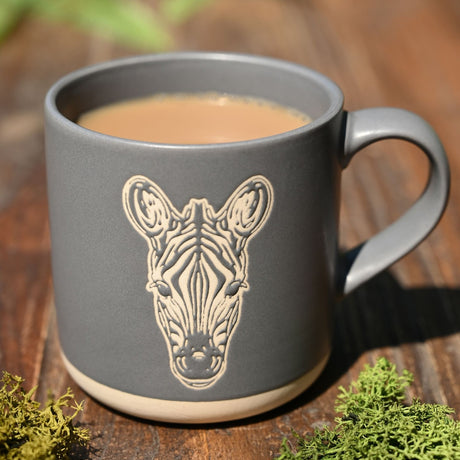 Grey stoneware mug with an engraved zebra head design in pale cream lines, a cream base band, and a curved handle, filled with tea on a wooden surface.
