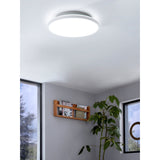 Round white LED downlight, Nelbenne 31.5cm, mounted flush on a ceiling in a grey room with a wooden shelf, plant, and windows below.