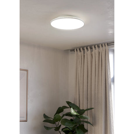 Nelbenne LED Downlight 50cm in white, a round flat ceiling fixture with a frosted diffuser, shown illuminated in a neutral-toned living room near curtains and a plant.