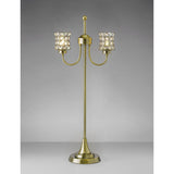 The Nelson 2 Light Crystal Table Lamp in antique brass features two arms, crystal-embellished shades, a round base, vertical stem, and decorative finial. It's shown against a plain gray background.