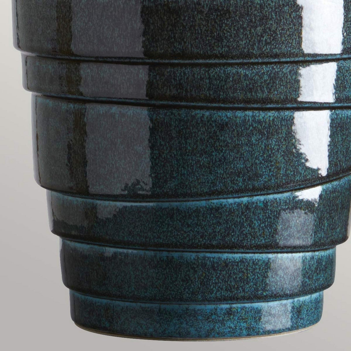 A close-up of a textured ceramic Neptune 1 Light Table Lamp showcases its deep teal glaze with horizontal ridges, capturing the vivid depth of a Blue Reactive Glaze. The lamp's surface reflects light, emphasizing its glossy finish, while the blurred background draws attention to the intricate details.