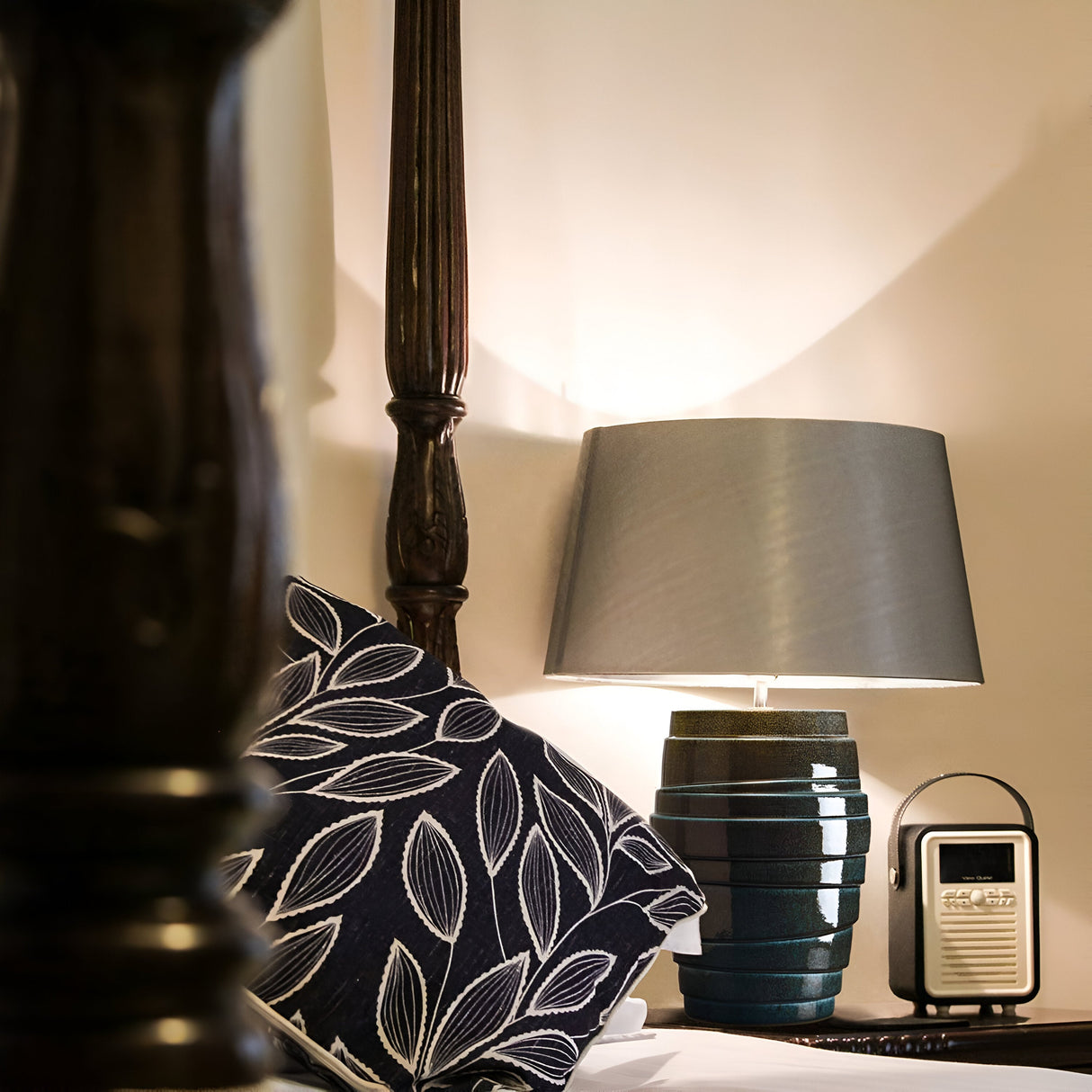 A cozy bedroom scene with a patterned pillow featuring leaf designs on a bed. Beside it, the Neptune 1 Light Table Lamp - Blue Glaze, adorned with polished nickel accents and blue reactive glaze, glows warmly. A retro-style radio sits next to the lamp on a wooden nightstand.