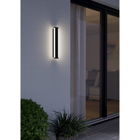 Neviano LED Outdoor Wall Light 38cm in black steel, mounted beside a glass door, glowing warm white along its vertical diffuser on an exterior house wall at night.
