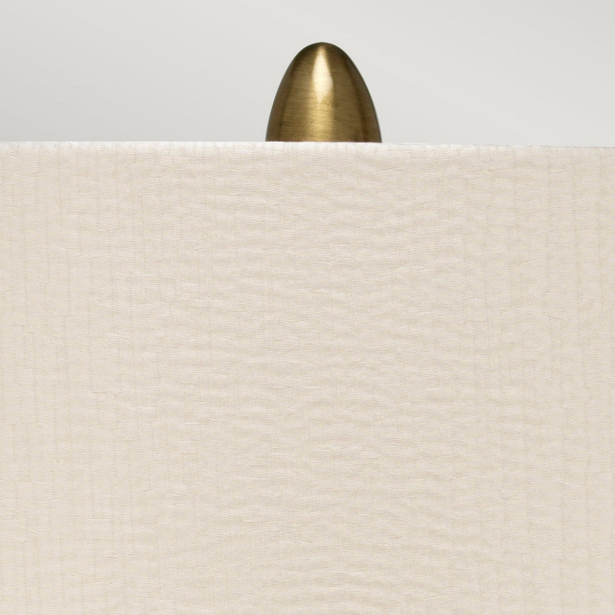 A brass, bullet-shaped finial peeks over the top edge of a Pearl Satin Shade, set against a neutral background. The Newham 1 Light Large Table Lamp - Bronze Ceramic/Pearl Shade showcases a cream-colored shade with a subtle textured pattern that elegantly complements its bronze ceramic base.