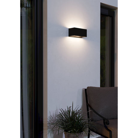 Nilvanne LED Outdoor Wall Light in black, rectangular aluminium body with clear glass, mounted on an exterior wall casting warm light upward and downward beside a patio seating area.