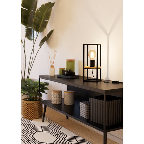 Ninarne table lamp with a black open steel cage frame, brown wooden centre shelf and exposed globe bulb, shown lit on a dark sideboard in a living room setting.