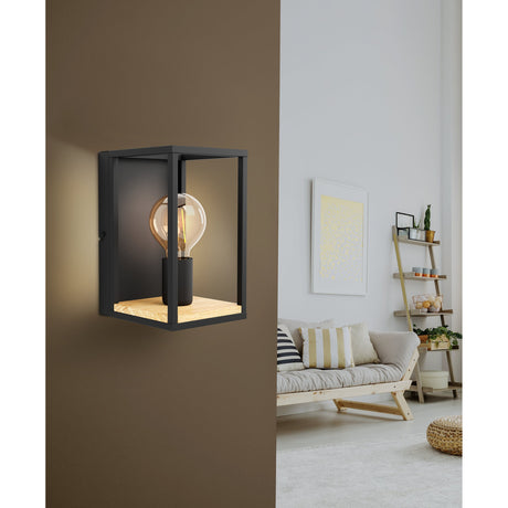 Ninarne wall light with black steel open box frame, wooden base, and upright bulb holder displaying a glowing globe bulb, mounted on a brown wall in a living room setting.