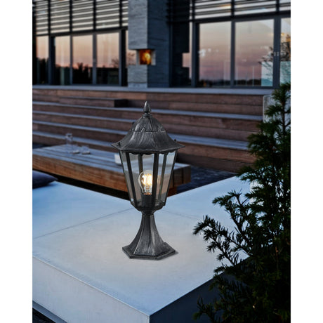 Nolmane Bollard Light in black and silver patina aluminium, hexagonal lantern head with clear glass panels and pointed finial on a pedestal base, shown illuminated on an outdoor terrace ledge.