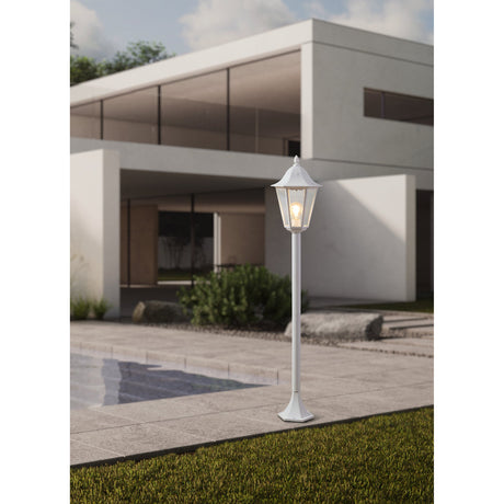 Nolmane Bollard Light in white, a traditional six-sided lantern post with clear glass panels and finial top, shown illuminated on a patio beside a modern house and pool.