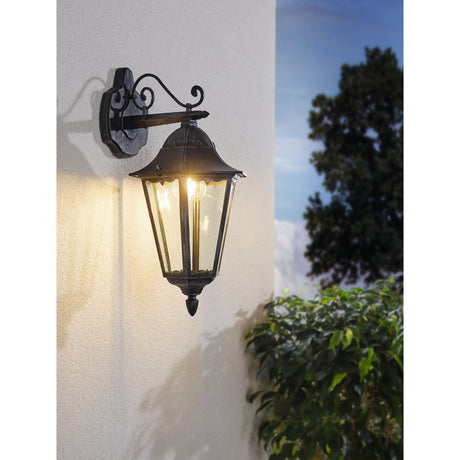 Nolmane downward outdoor wall light, a six-sided black and silver patina lantern on a scrolled arm, mounted on a light wall beside greenery and trees.