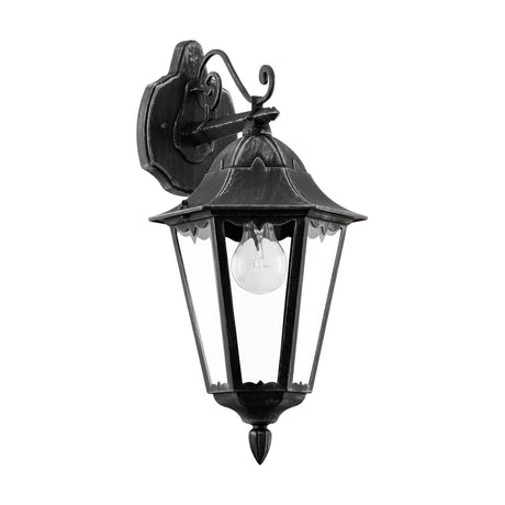 Nolmane downward outdoor wall light in black with silver patina, a six-sided lantern with clear glass panels on a scrolled arm and shaped wall plate, shown on a white background.