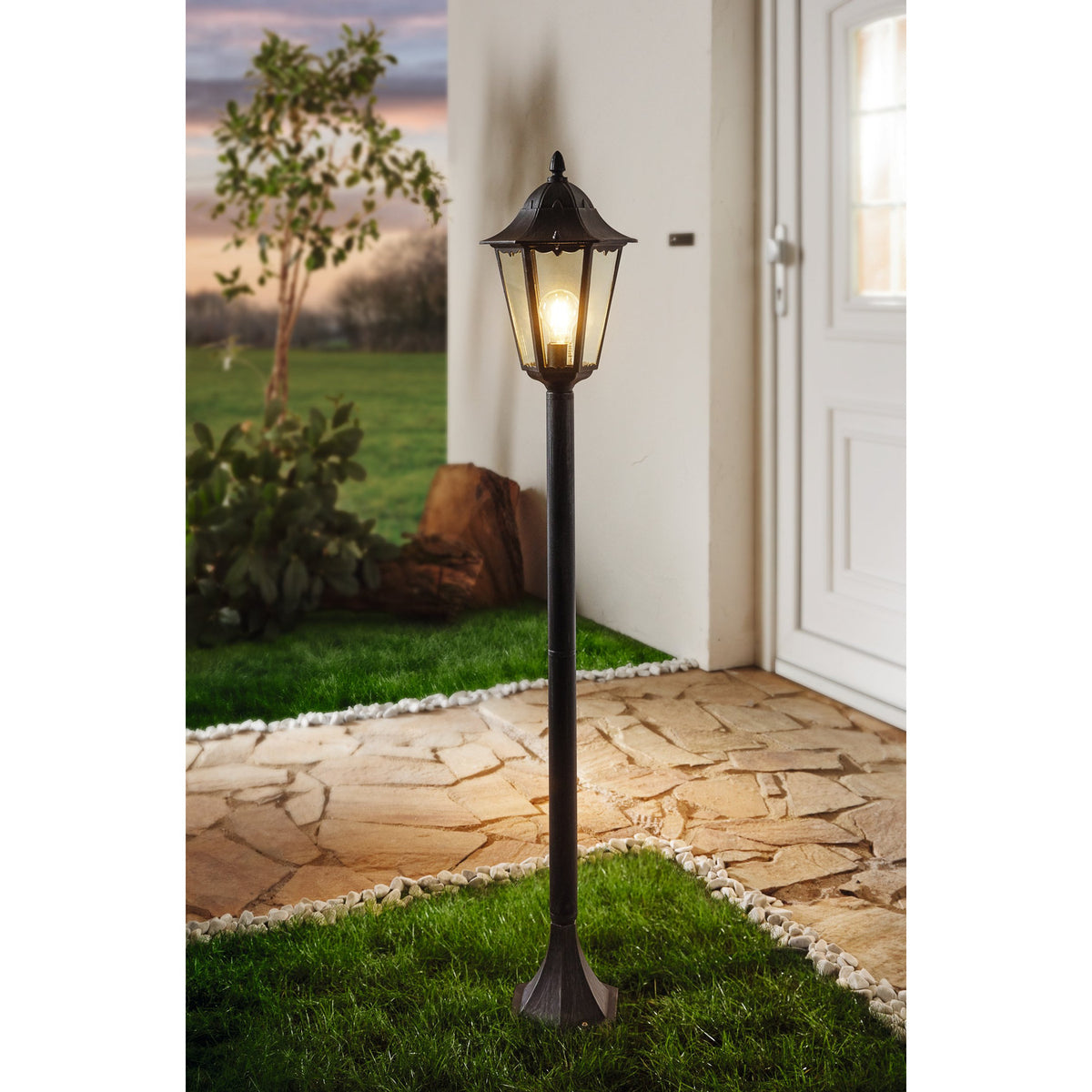 Nolmane outdoor floor lamp with hexagonal glass lantern head and pointed finial on a slim black aluminium post, illuminated beside a house entrance on a stone path.