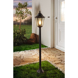 Nolmane outdoor floor lamp with hexagonal glass lantern head and pointed finial on a slim black aluminium post, illuminated beside a house entrance on a stone path.