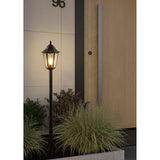 Nolmane outdoor floor lamp, a black and silver patina Victorian-style lantern with hexagonal glass head on a slim post, lit beside a planted entrance and wooden door.