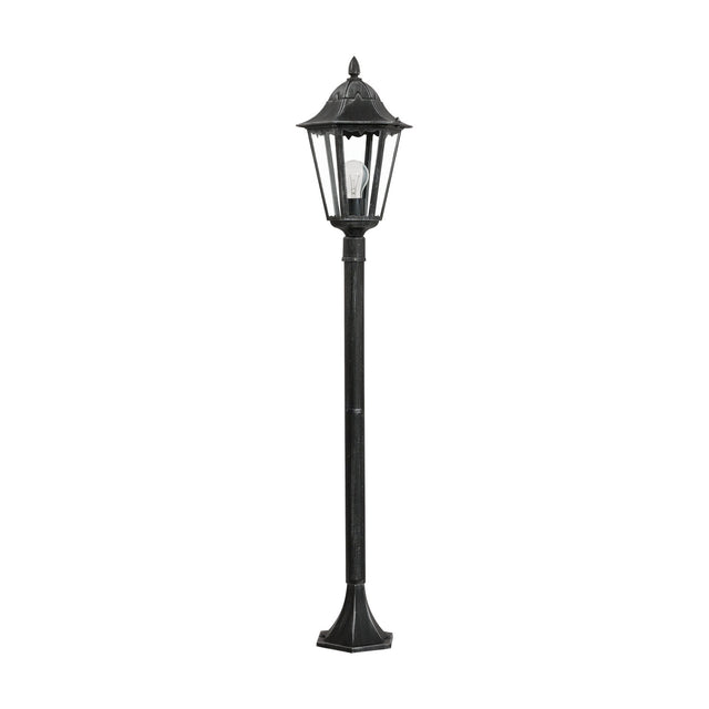 Nolmane outdoor floor lamp with a hexagonal glass-panelled lantern head, pointed finial, and slim post on a flared base, in black cast aluminium with silver patina, shown on a plain white background.