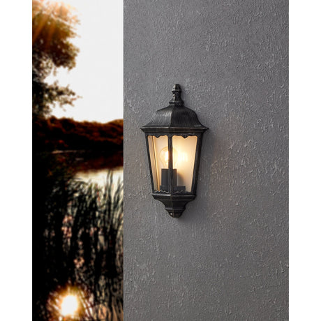 Nolmane outdoor wall light with a lantern-style aluminium body in black with silver patina, domed roof, finial, and clear glass panes, mounted on a grey textured wall.