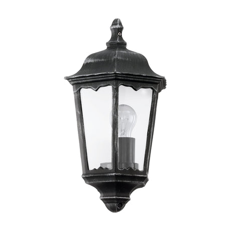 Nolmane half-lantern outdoor wall light with black and silver patina cast aluminium body, domed roof, finial top, and clear glass panes showing an E27 bulb, on a white background.