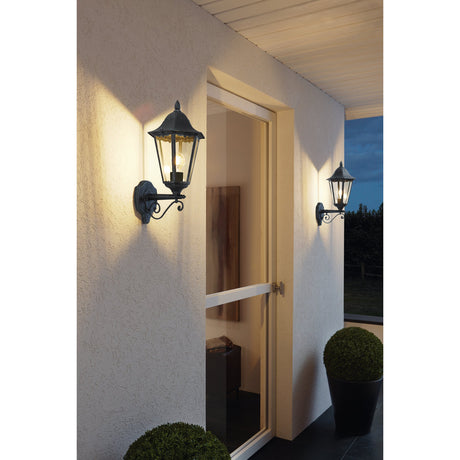 Nolmane upward outdoor wall light in black with silver patina, hexagonal lantern on a scroll bracket, shown illuminated beside a doorway on a house exterior at dusk.