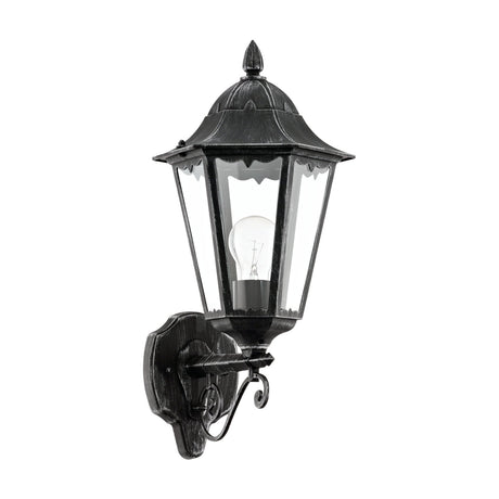 Nolmane upward outdoor wall light in black with silver patina, hexagonal lantern with clear glass panels on a cast aluminium scroll bracket, shown with E27 bulb on a plain white background.