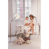 Two young girls sit on a white vintage desk by a tall window, reading together. Colorful pillows, books, and toys are scattered nearby, softly illuminated by the Nordica 1 Light Floor Lamp in Matt White with an Ivory White Shade.