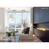 A modern living room with large windows, city view, beige sofa, Nordica 1 Light Floor Lamp - Matt White With Ivory White Shade, tall vase with branches, indoor plants, and a long built-in fireplace on a dark accent wall.