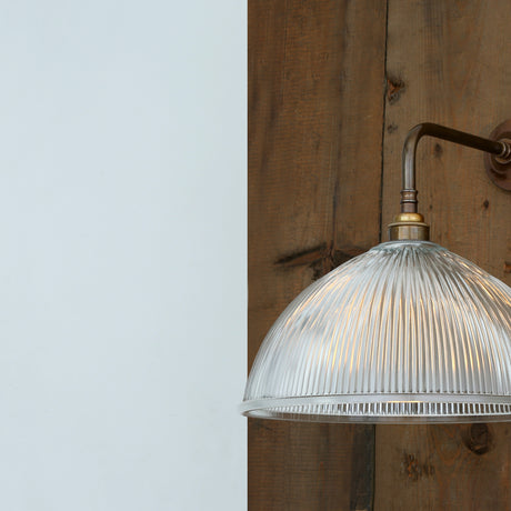 Nova Ribbed Glass Wall Light with a domed, vertically ribbed glass shade on a curved brass arm and round wall backplate, mounted on wood against a white wall.