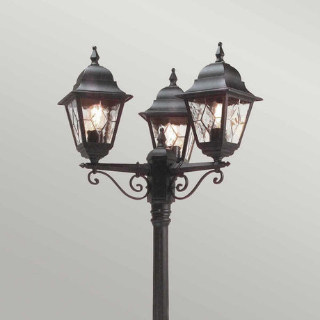 The Norfolk 3 Light Lamp Post - Black stands against a plain background, showcasing a decorative design with three lantern-style lights encased in clear glass panels. This traditional lamp post boasts ornate metalwork and finials on top, adding an elegant and classic charm to outdoor lighting.