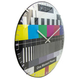 Round 35cm wall clock with domed glass face showing a TV test card design in bold yellow, cyan, green, magenta and red bars, black grid lines and slim silver hands, on a white background.