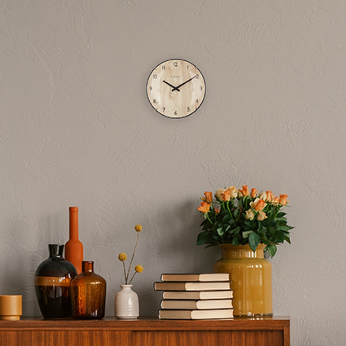 Edge Wood Mini Wall Clock 20cm, round pale wood face with black numerals and hands, slim black rim, mounted on a grey wall above a sideboard with vases, books and flowers.
