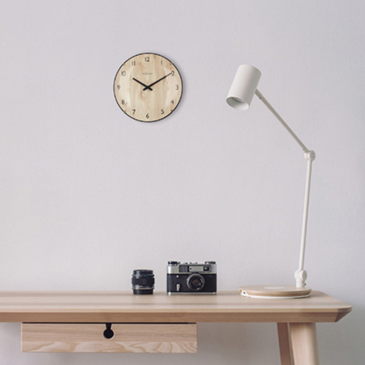 Edge Wood Mini Wall Clock, 20cm round, light wood face with black numerals and slim black rim, mounted above a desk with a lamp and camera.