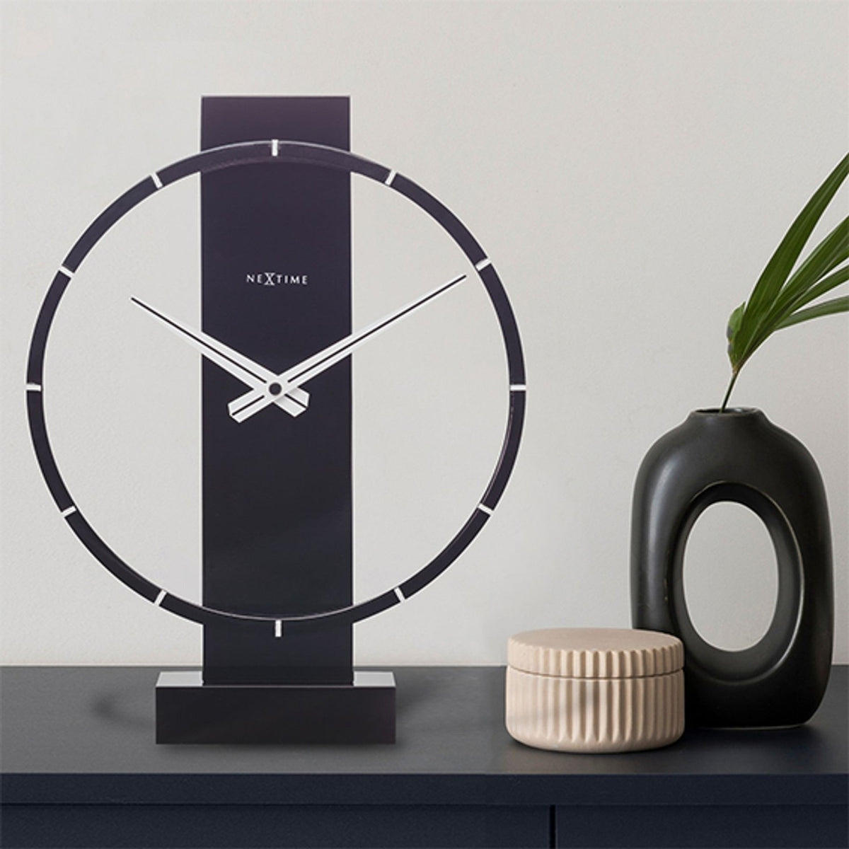Black 34cm open-ring wood and steel table or wall clock with white baton markers and slim hands, shown on a dark sideboard beside a vase and trinket box.