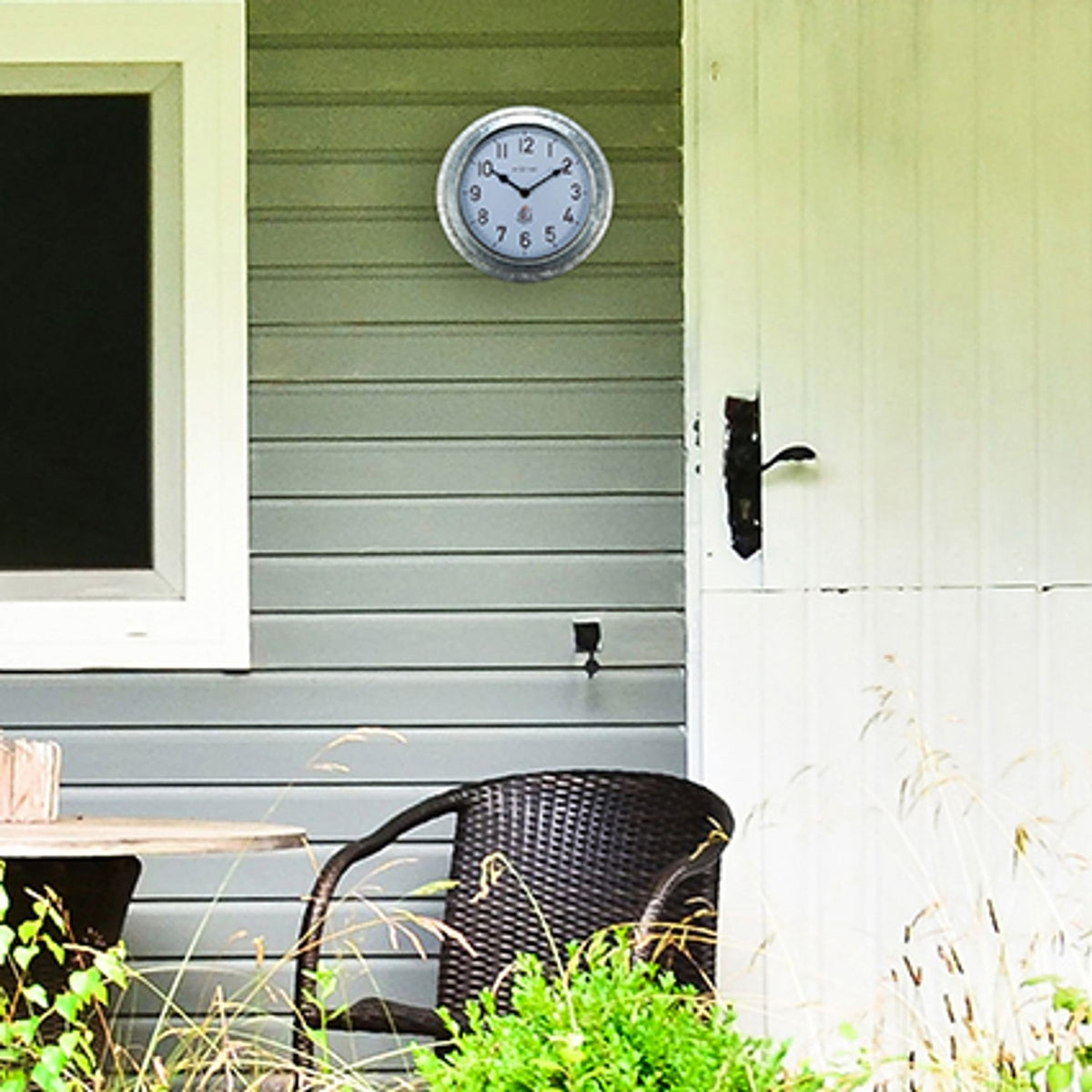 Poppy Galvanised Colour Deep Round Wall Clock 22cm in silver with white dial and black hands, mounted on grey porch siding beside a door, wicker chair below.
