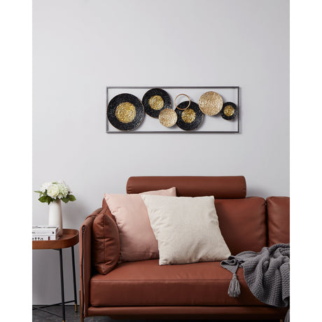 Numazu metal wall decoration in black and gold, a slim rectangular frame holding overlapping textured discs and a ring, shown above a brown leather sofa with cushions.