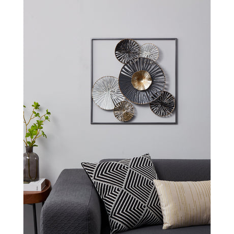 Numazu square metal wall decoration in a slim dark frame, featuring overlapping black, white and gold radiating discs with a ridged gold centrepiece, shown above a sofa with patterned cushions.