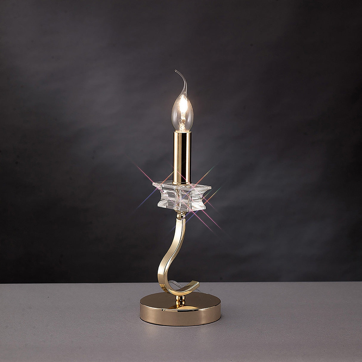 The Nydia 1 Light Crystal Table Lamp in French gold features a star-shaped crystal accent and a round base, displayed on a gray surface against a dark background.