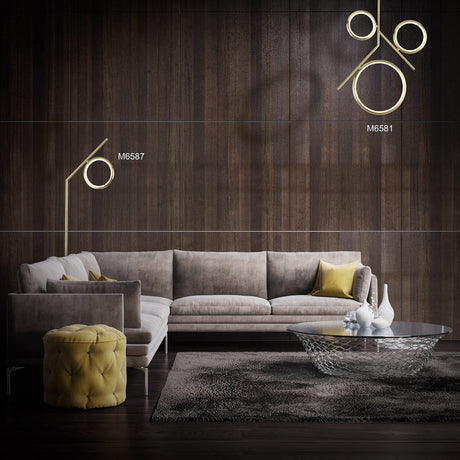 A modern living room with a gray sectional sofa, yellow cushions, a round tufted ottoman, an Olimpia LED Floor Lamp 12W 3000K in satin gold against a dark wood wall, a glass coffee table, and white vases on a gray rug.