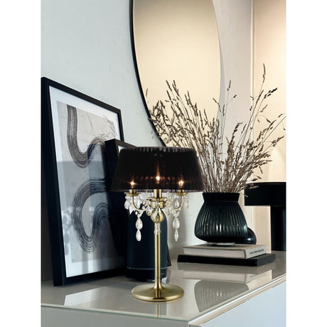 A modern console table displays the Olivia 3 Light Crystal Table Lamp in antique brass with a black shade, abstract art, books, a black vase with dried grass, and is topped with a round wall mirror for a chic and timeless appeal.