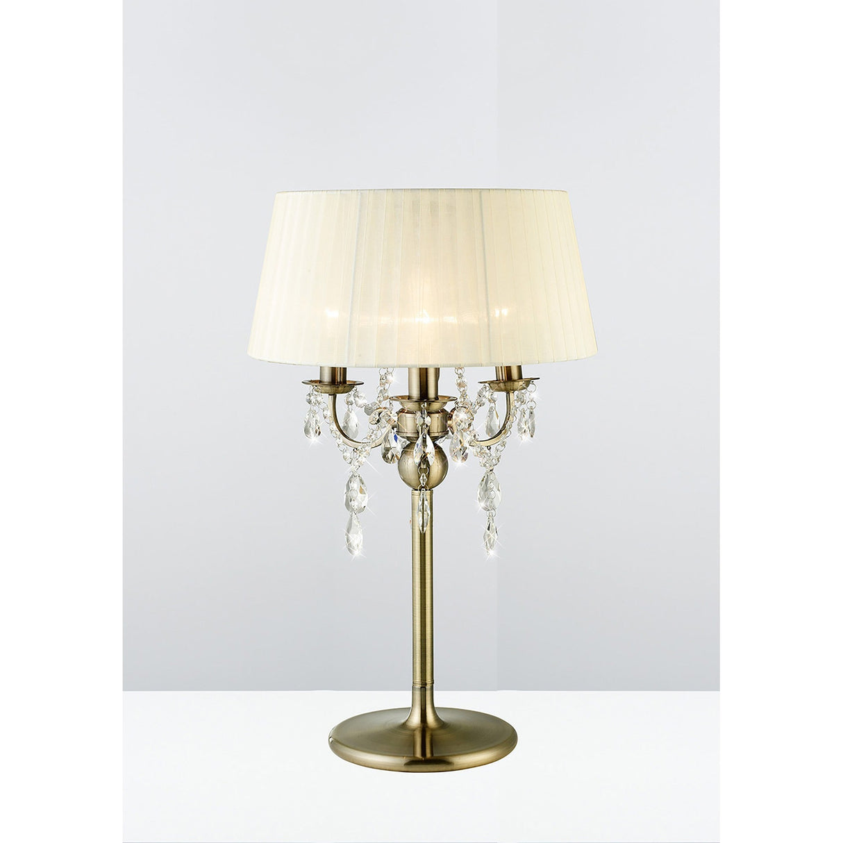 The Olivia 3 Light Crystal Table Lamp in antique brass features a round base, cream pleated shade, and crystal accents on curved arms, shown against a plain light background.