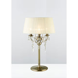 The Olivia 3 Light Crystal Table Lamp in antique brass features a round base, cream pleated shade, and crystal accents on curved arms, shown against a plain light background.